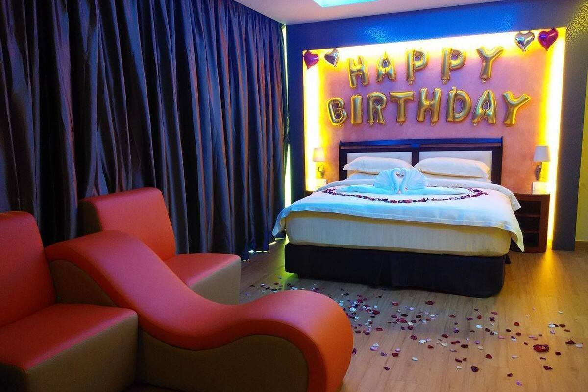 A hotel room with a bed, a red sofa, and a 'Happy Birthday' balloon decoration.