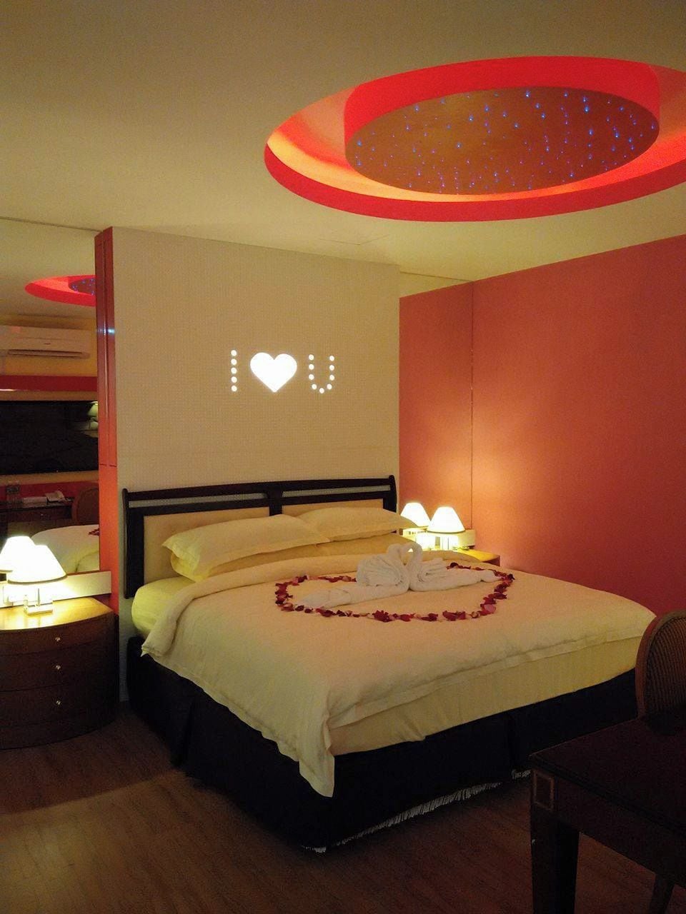 A hotel room with a bed, two bedside lamps, a wall with a heart-shaped light, and a ceiling light fixture.