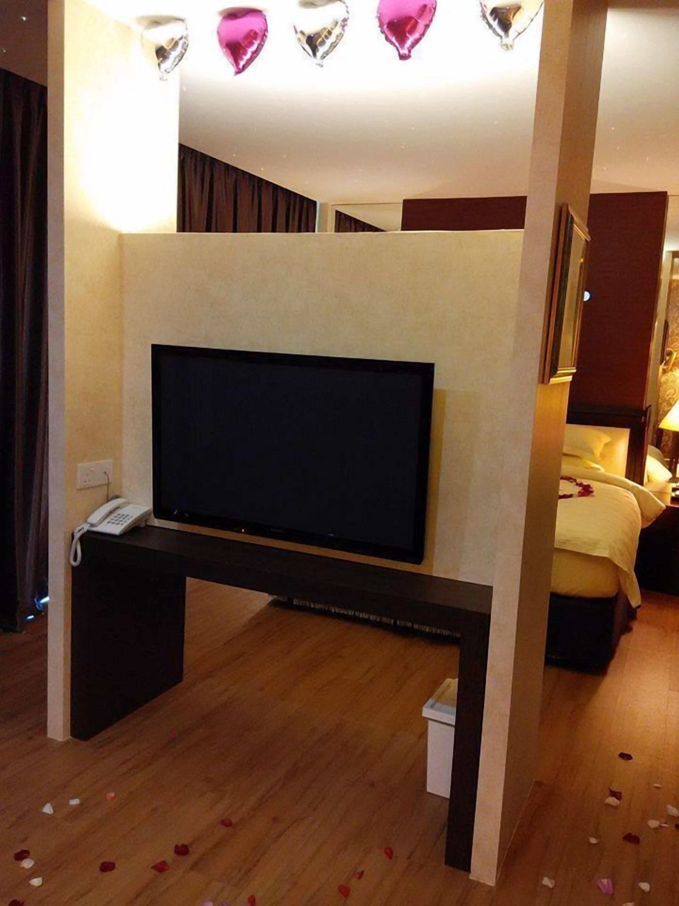 A hotel room with a flat-screen TV on a stand, a bedside table with a phone, and a bed with a floral bedspread.