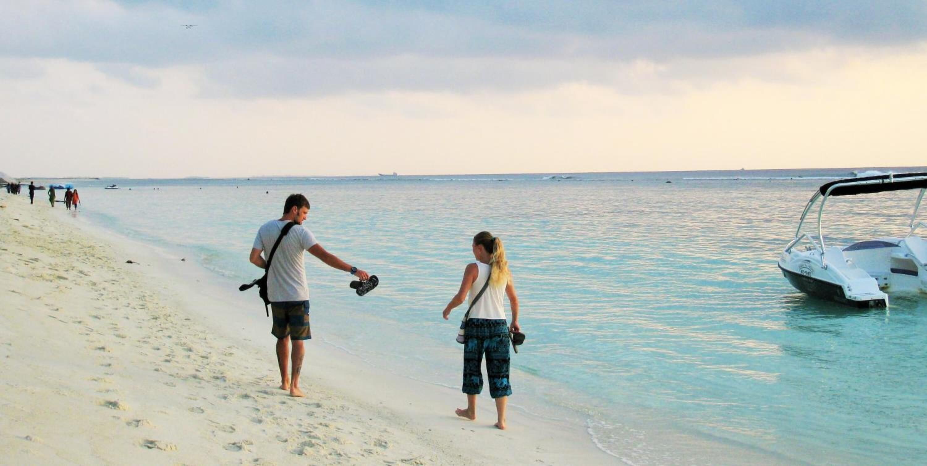 Escena de playa con un bote amarrado a la derecha, gente caminando por la orilla y un cielo despejado.