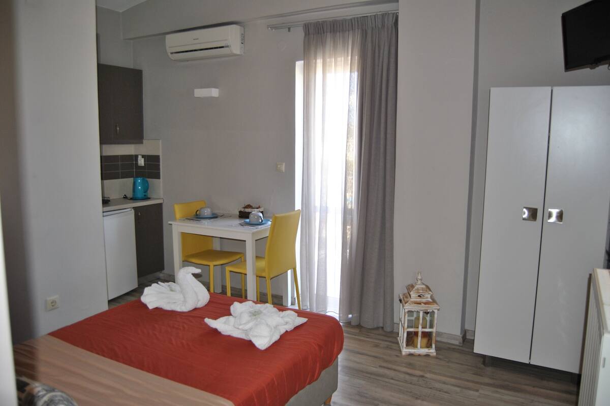 A hotel room with a bed, a desk, a chair, a small table, a white cabinet, and a window with curtains.