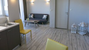 A modern living room with a sofa, a coffee table, and a dining area with a chair and a small table. There is a door and a window with curtains. - Pantheon Apartments (Chania)