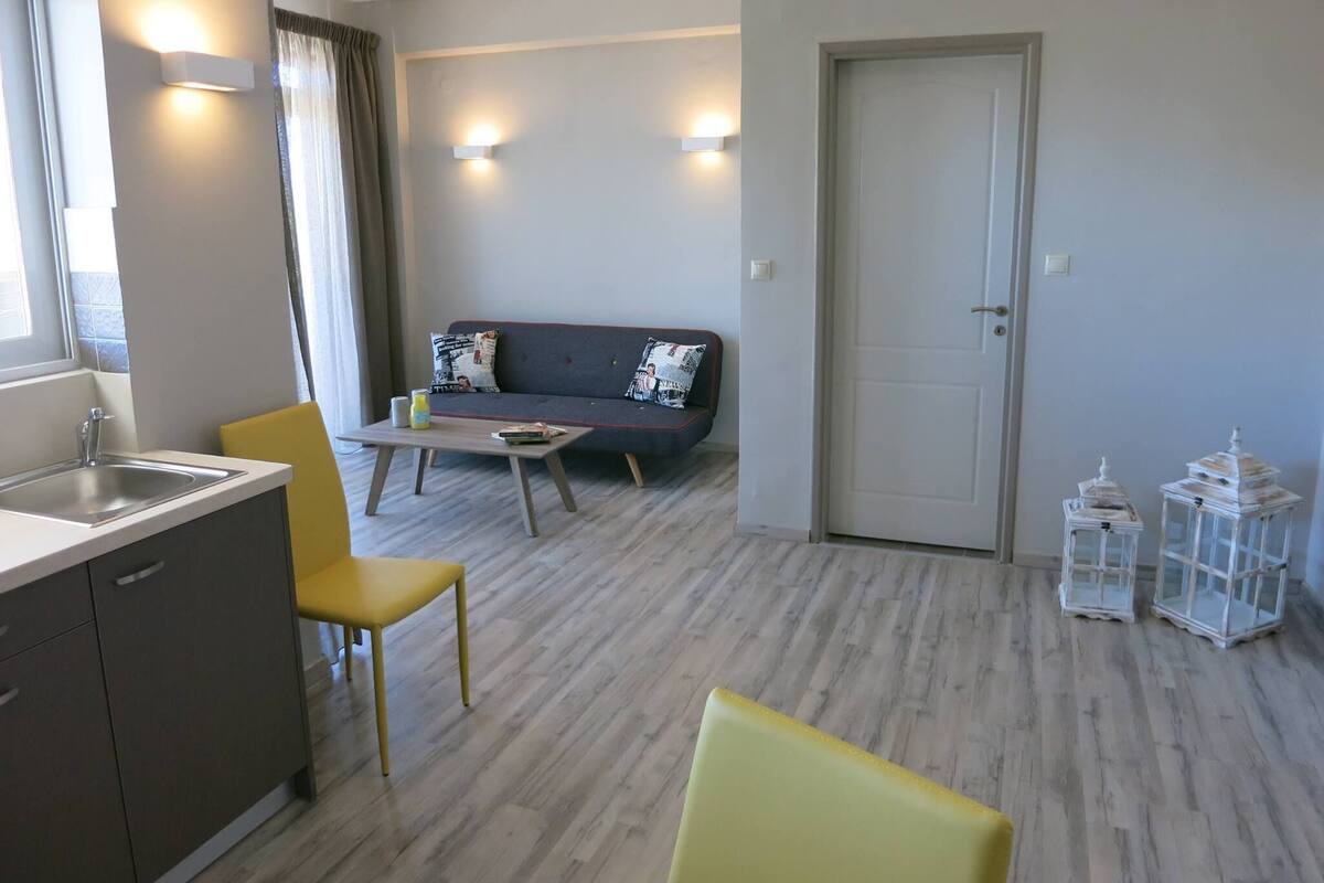 A modern living room with a sofa, a coffee table, and a dining area with a chair and a small table. There is a door and a window with curtains.