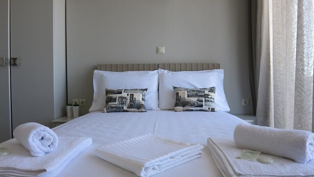 A neatly made bed with two pillows, a bedside table with a lamp, and a window with curtains. - Pantheon Apartments (Chania)
