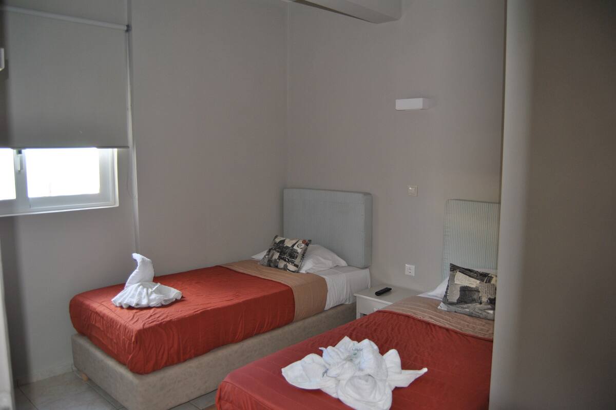 A hotel room with two beds, each with a red bedspread and a white pillow. There is a nightstand with a remote control and a lamp on it.