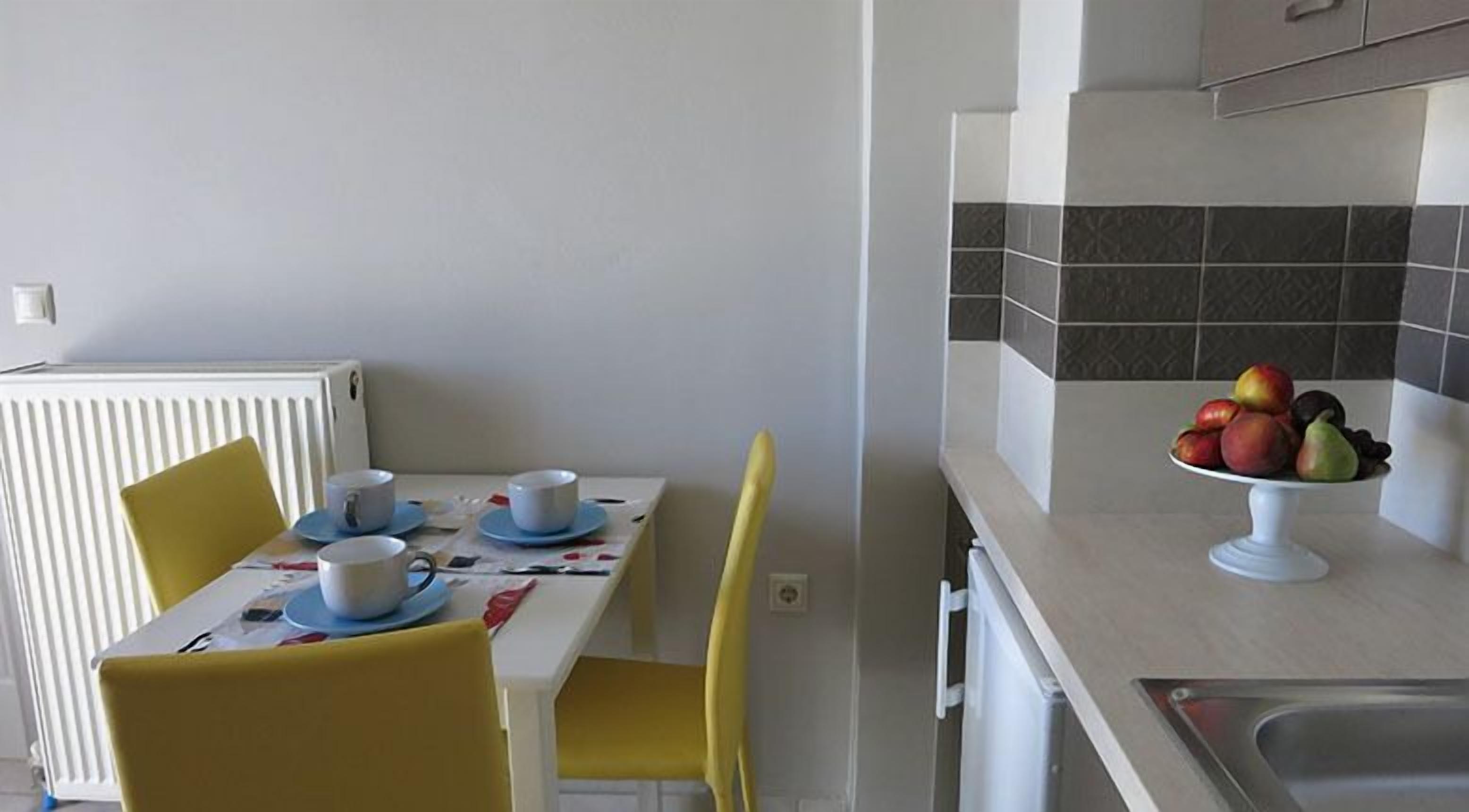 A dining area with a table set for two, yellow chairs, and a fruit bowl on the counter.