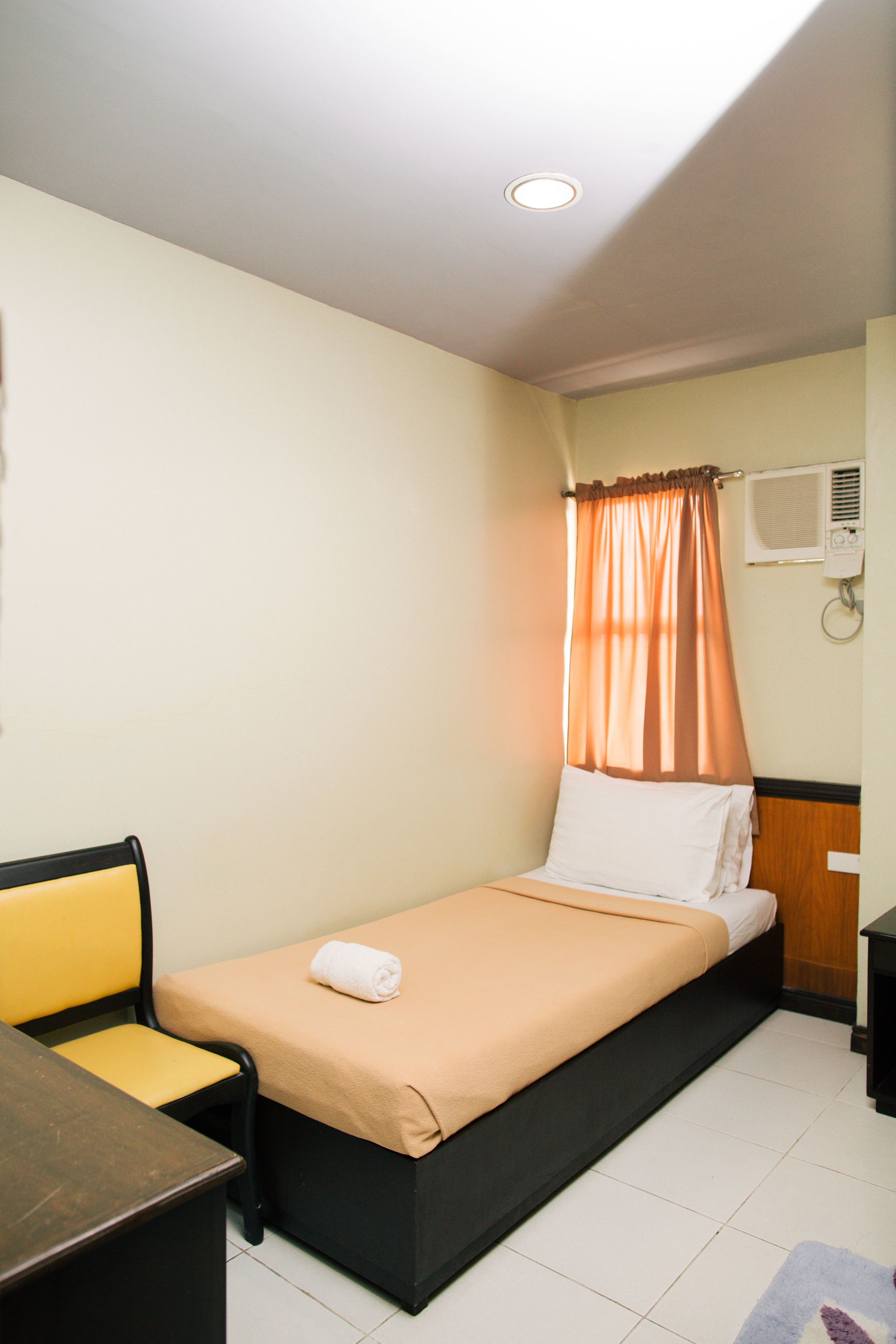 A single bed with a beige bedspread, a white pillow, and an orange curtain. There is a wooden nightstand to the right of the bed, and a yellow chair with a black frame is positioned to the left.