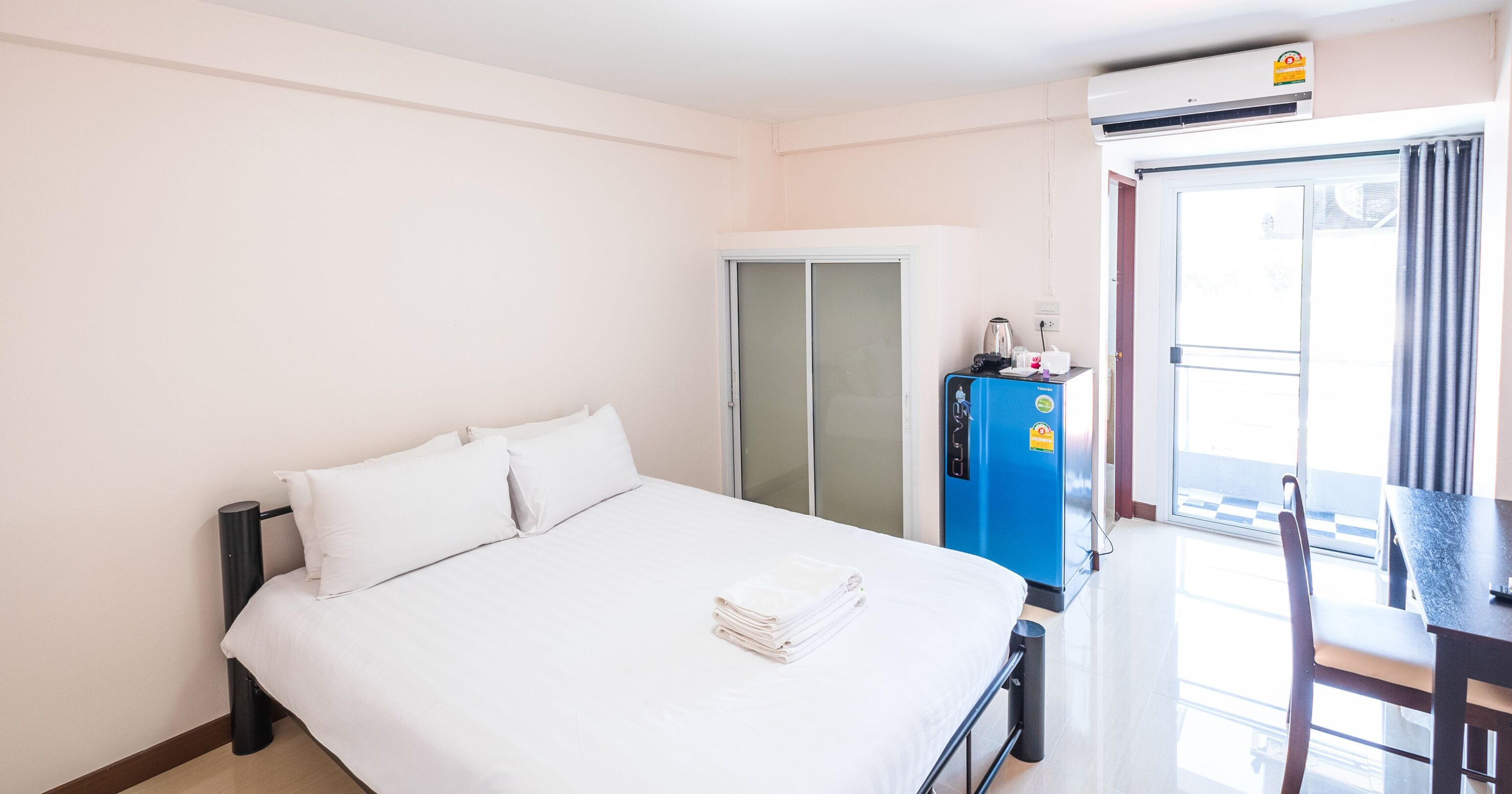 A hotel room with a bed, a wardrobe, a desk with a chair, and an air conditioning unit.