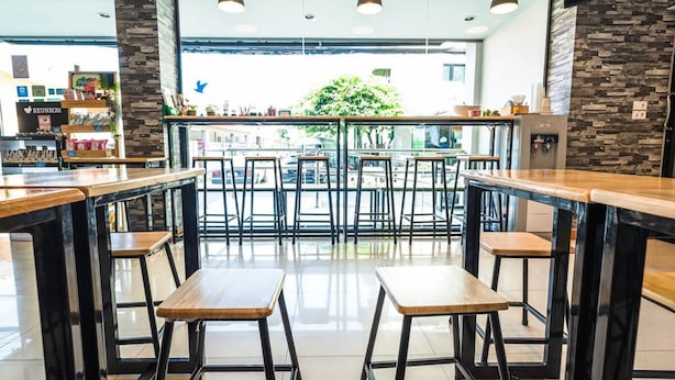 A modern cafe with wooden tables and black metal chairs, large windows, and a view of the street outside. - Don Muang Hotel (Bangkok)