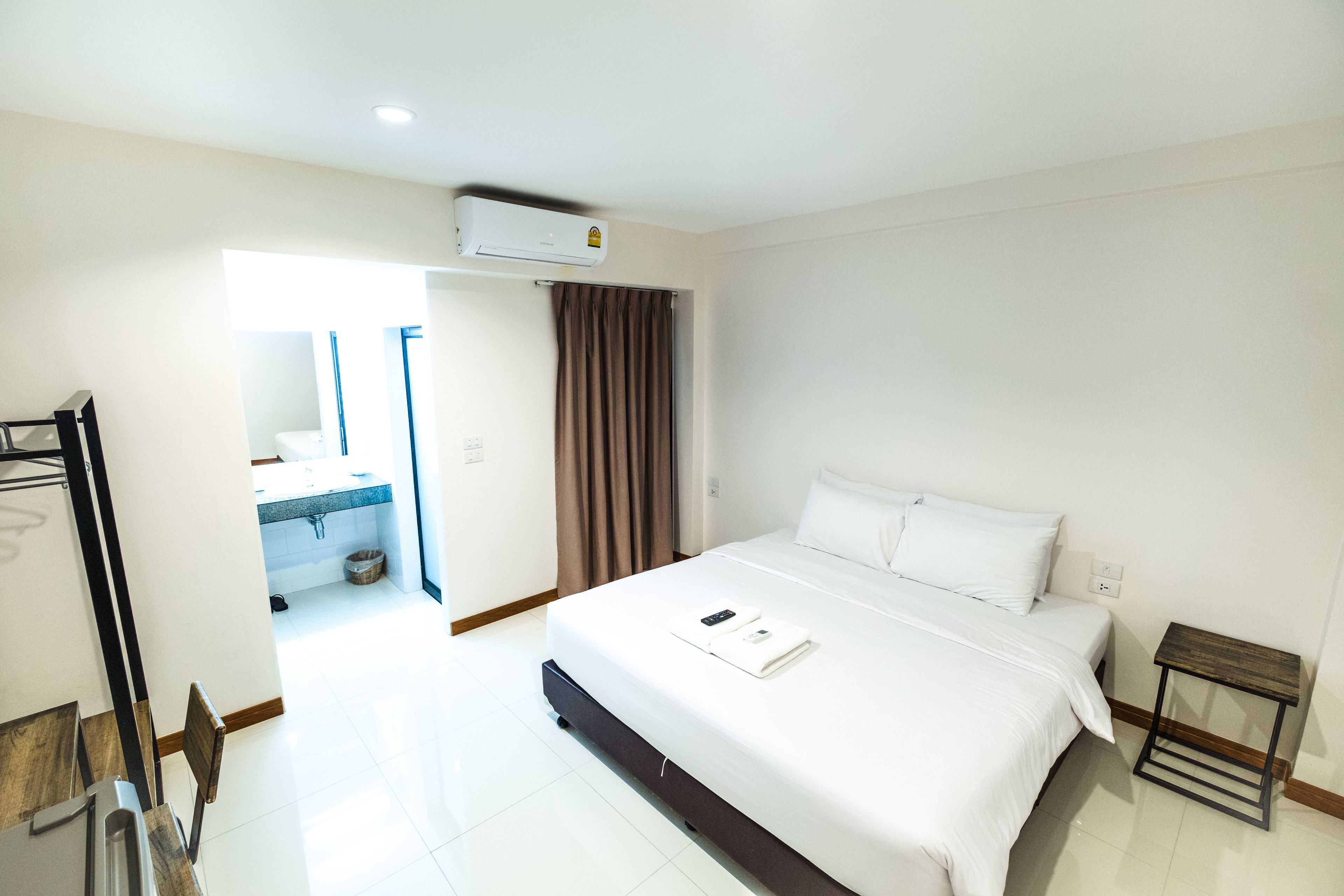 A hotel room with a bed, a small table, a mirror, and an air conditioning unit.