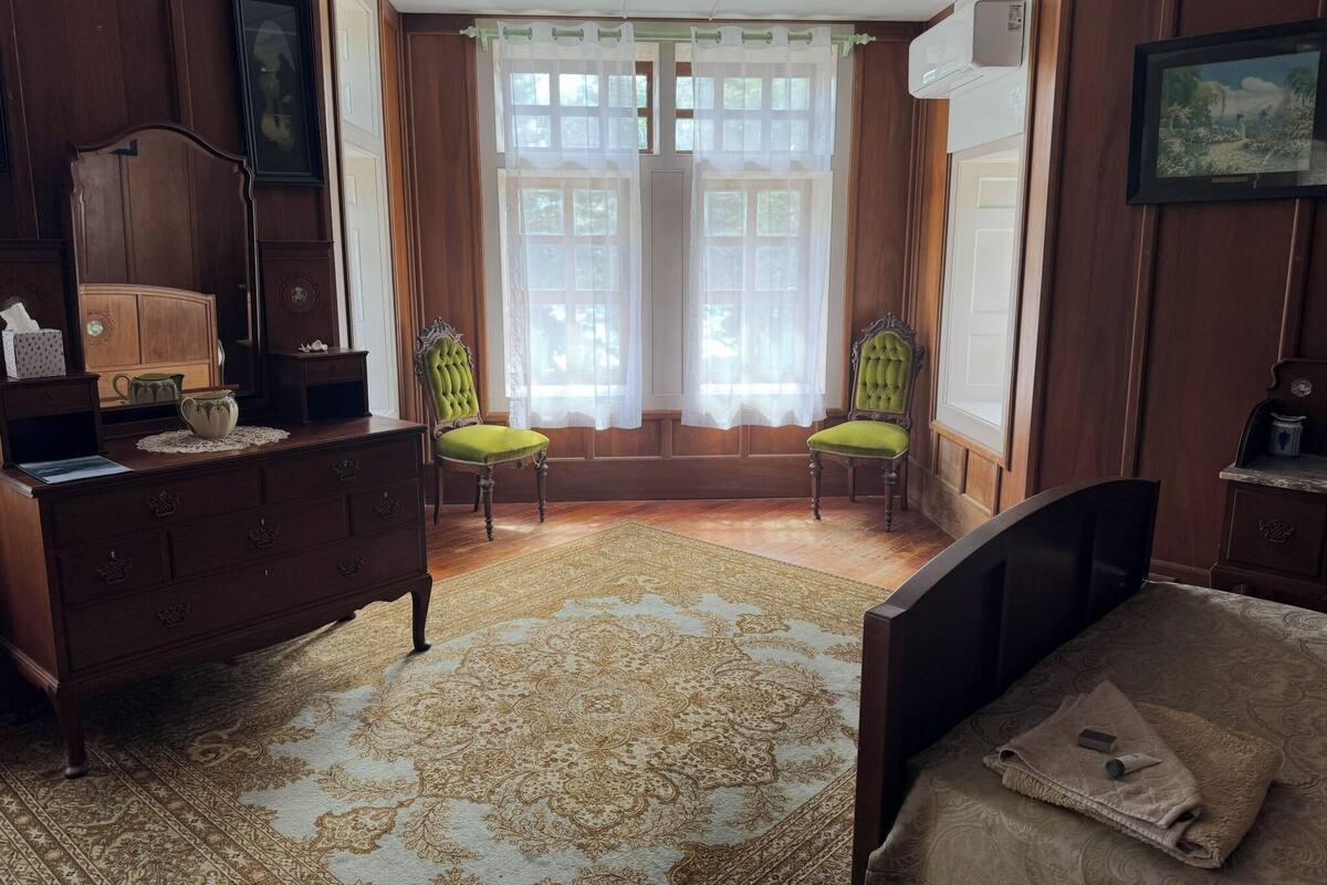 A room with a wooden dresser, two green upholstered chairs, a patterned rug, a bed with a brown blanket, and a window with sheer curtains.