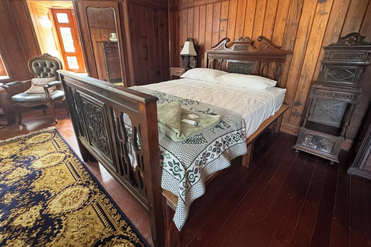 A wooden bed with a patterned bedspread, a nightstand with a lamp, and a vintage armoire in a room with wooden walls and floors.
