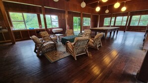 A spacious room with wooden flooring, large windows, wicker furniture, and hanging lights. - Oceania House (Home Island)