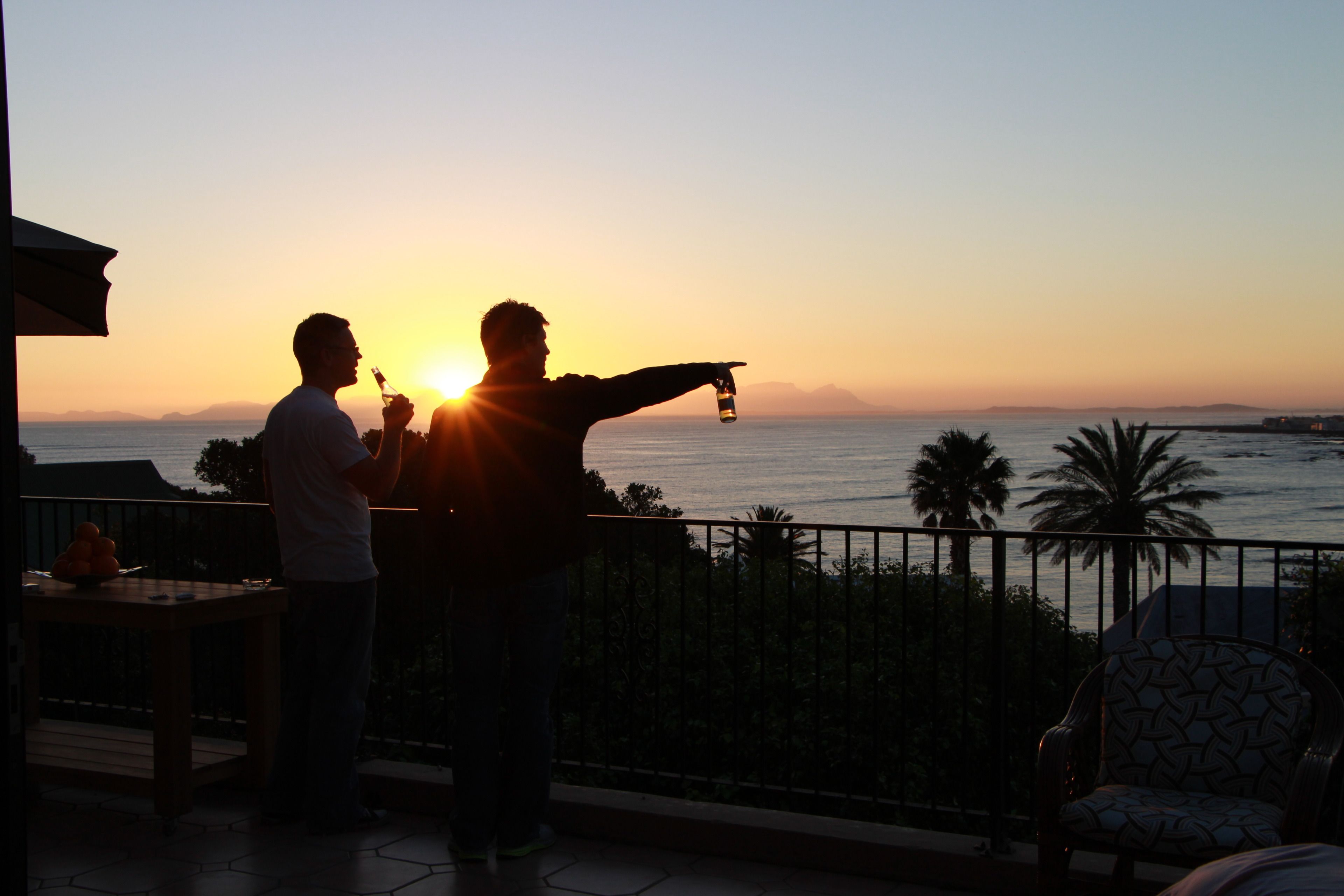 A balcony with a view of the sunset over the sea, a table with fruits, and a chair.
