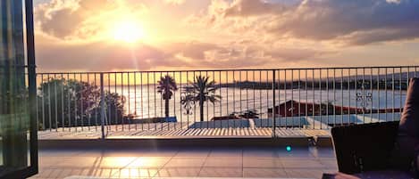 A balcony with a glass table, a cushion, and a view of the sunset over the sea.