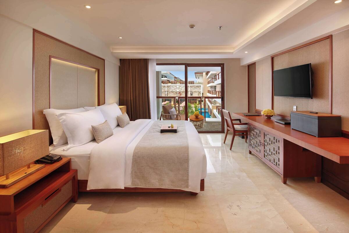 Habitación de hotel con una cama grande, un escritorio con televisor y un balcón con vistas a otros edificios.