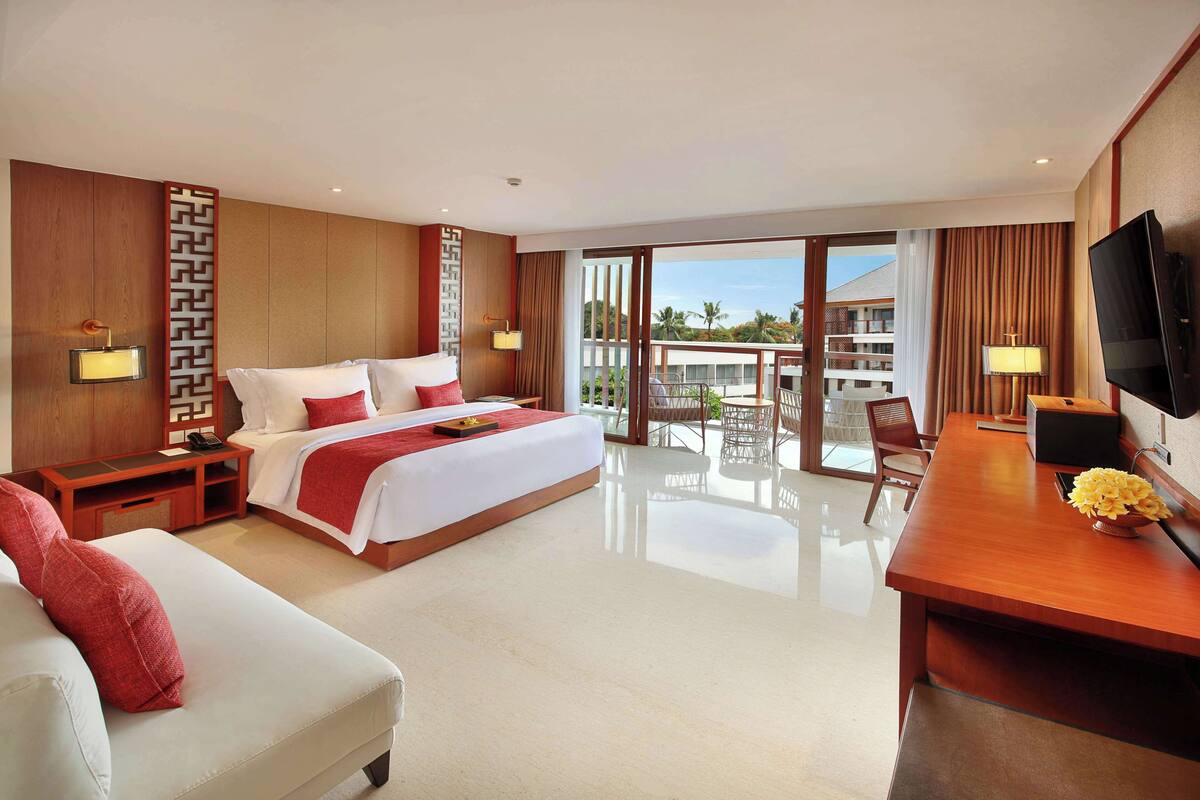 Habitación de hotel moderna con una cama grande, un escritorio, un televisor y un balcón con vistas a palmeras.