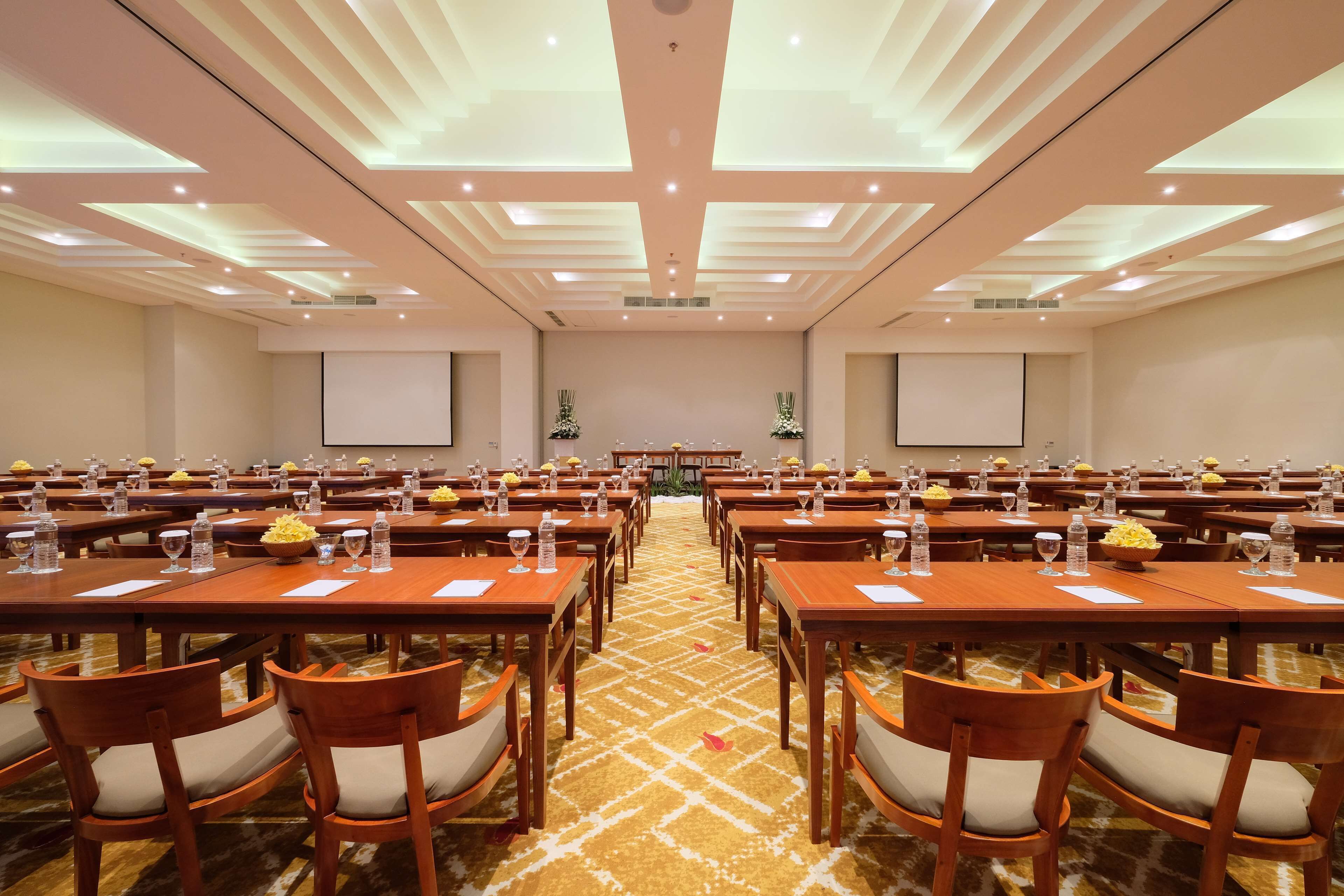 A conference room with multiple tables and chairs, equipped with water bottles and glasses, and a large screen at the front.