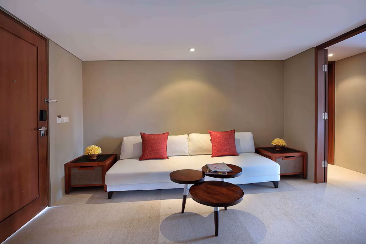 A modern hotel room with a white sofa, two red pillows, a small wooden side table with a vase, and a round coffee table.