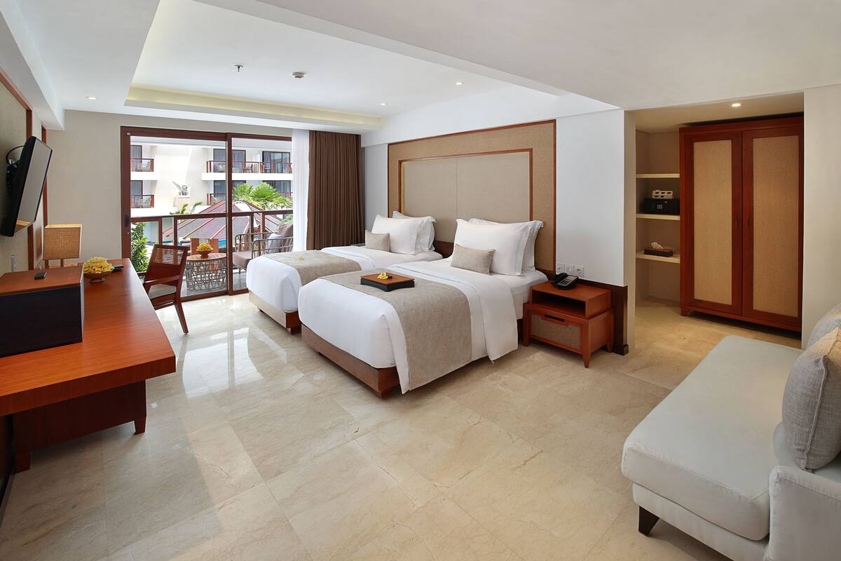 Habitación de hotel con dos camas, un escritorio, una silla, un armario y un balcón con vista al edificio.