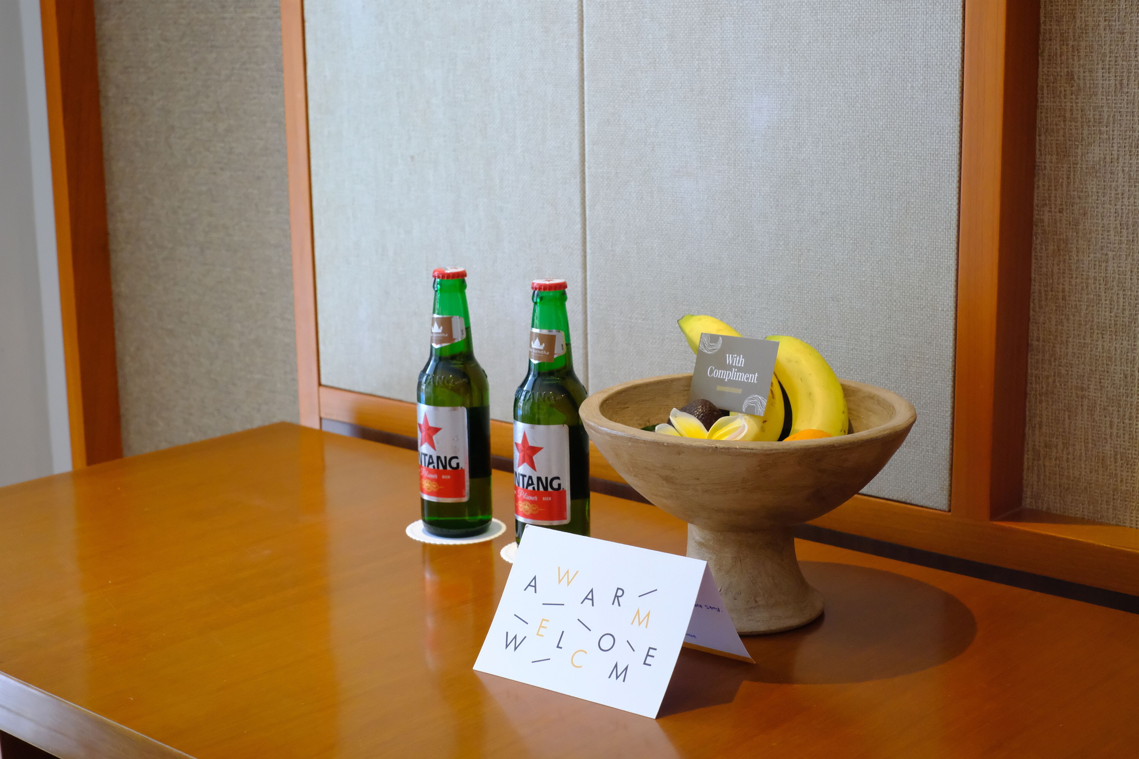 A wooden table with two bottles of beer, a bowl of fruit, and a welcome card.