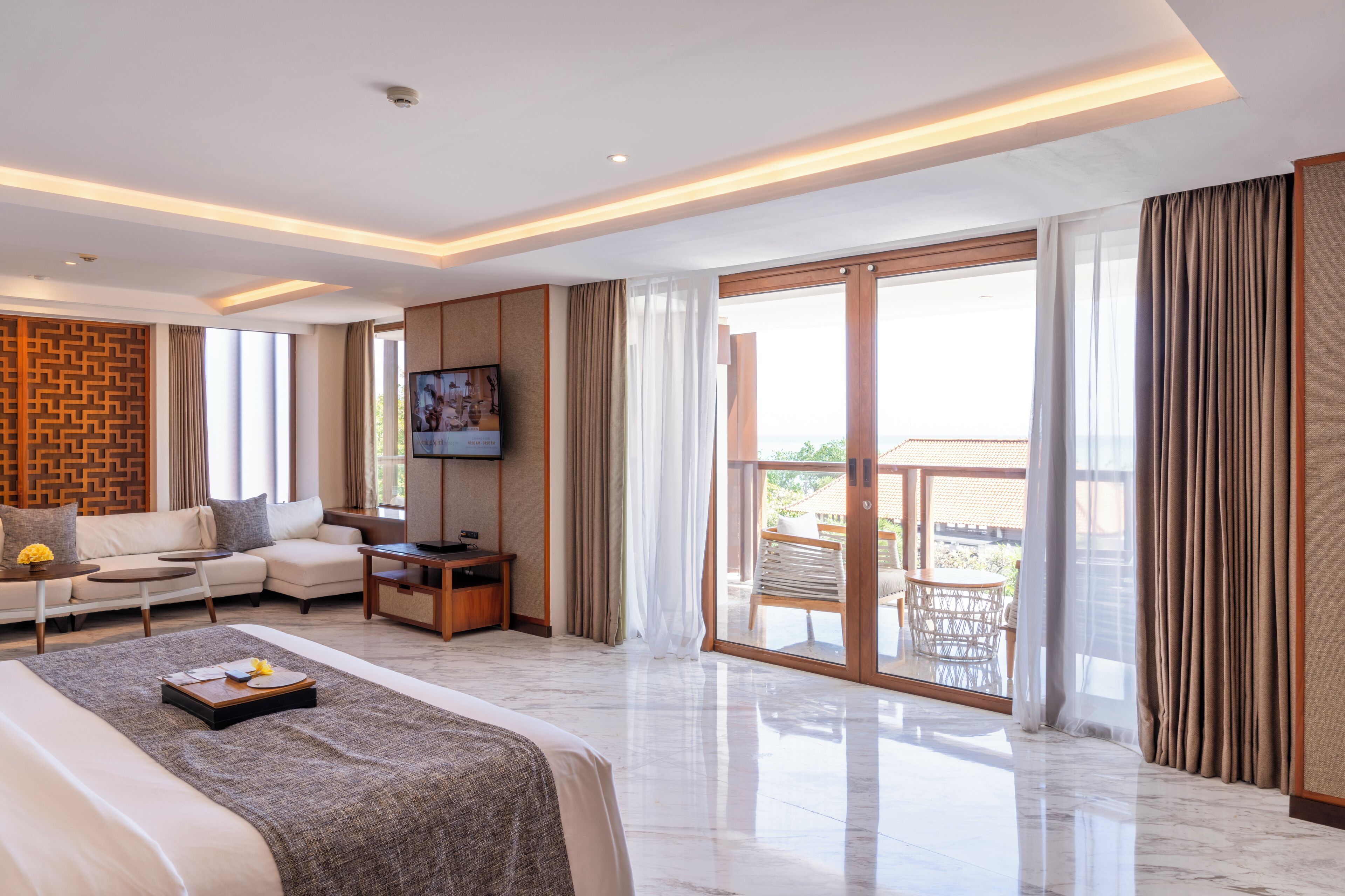 A modern hotel room with a large bed, a sofa, a TV, and a balcony with outdoor furniture.
