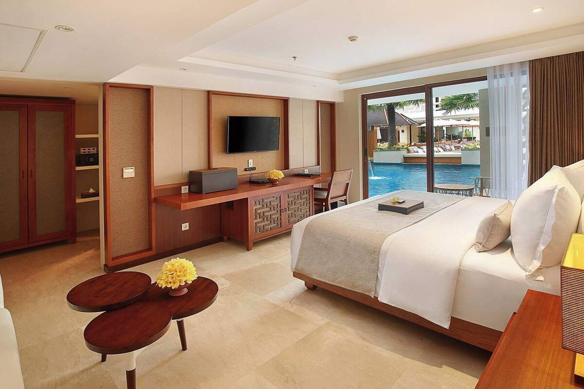 Habitación de hotel con cama, escritorio con televisor, una mesita con flores y vista a una piscina.