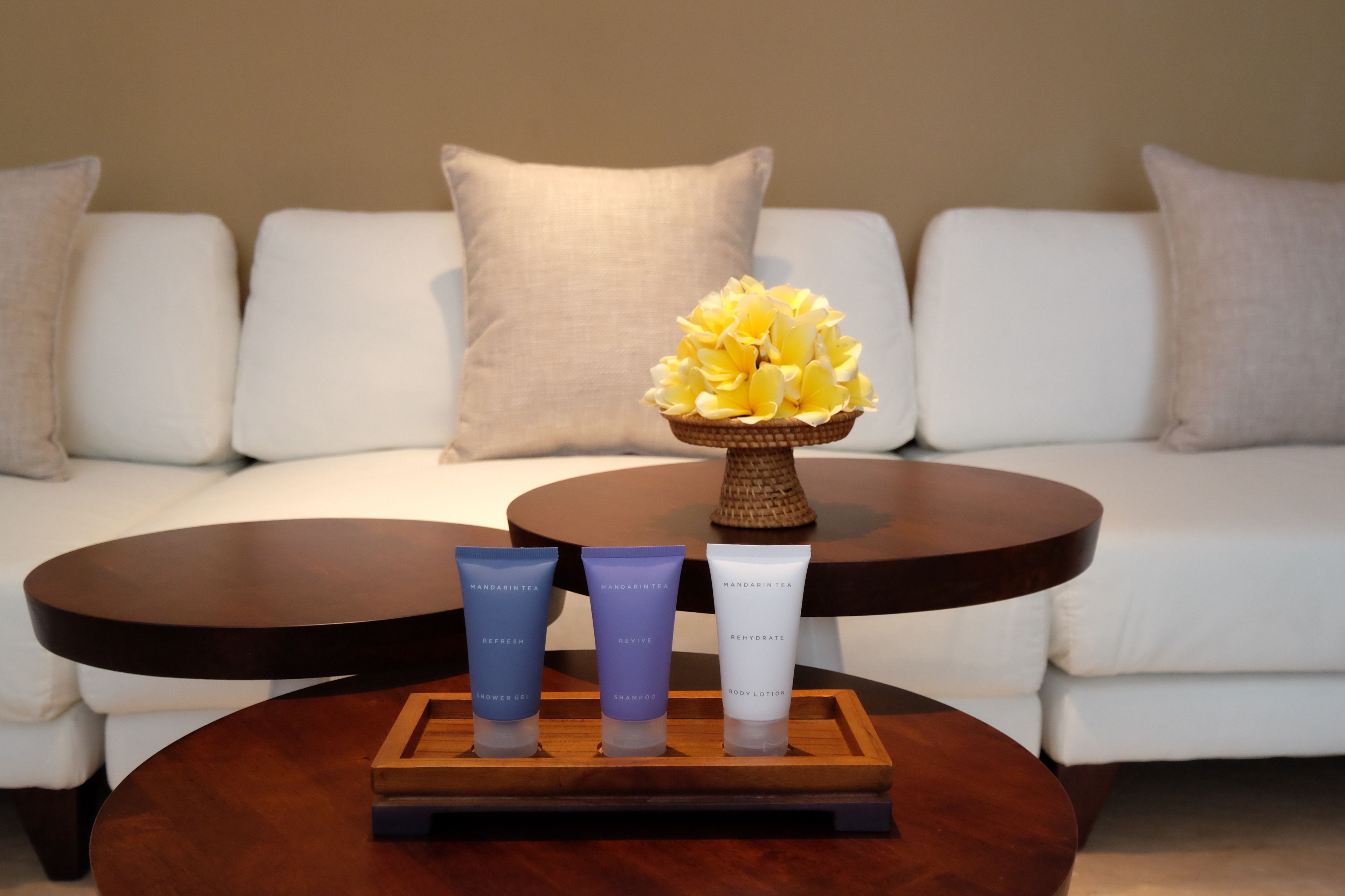 A white sofa with a wooden coffee table in front, holding three bottles of Mandarin Tea products and a vase with yellow flowers.