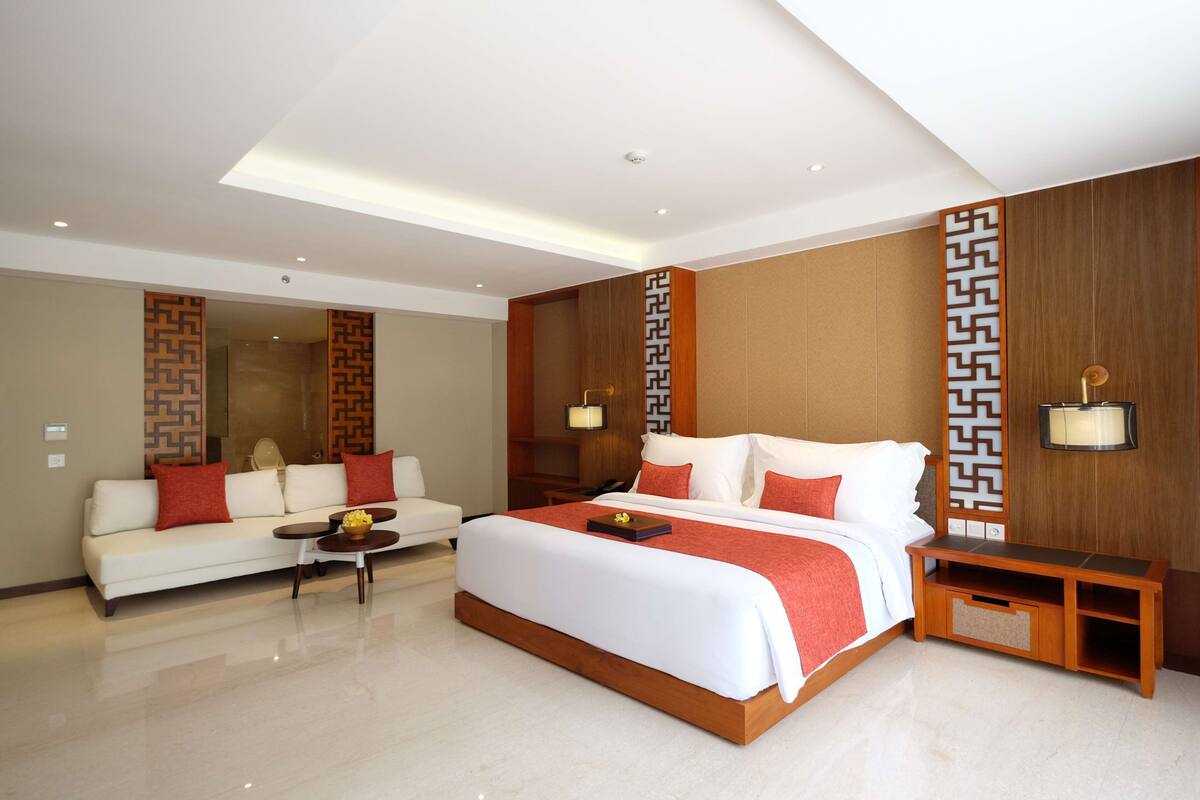 Una habitación de hotel moderna con una cama grande, un sofá, una mesita pequeña y una mesita de noche.
