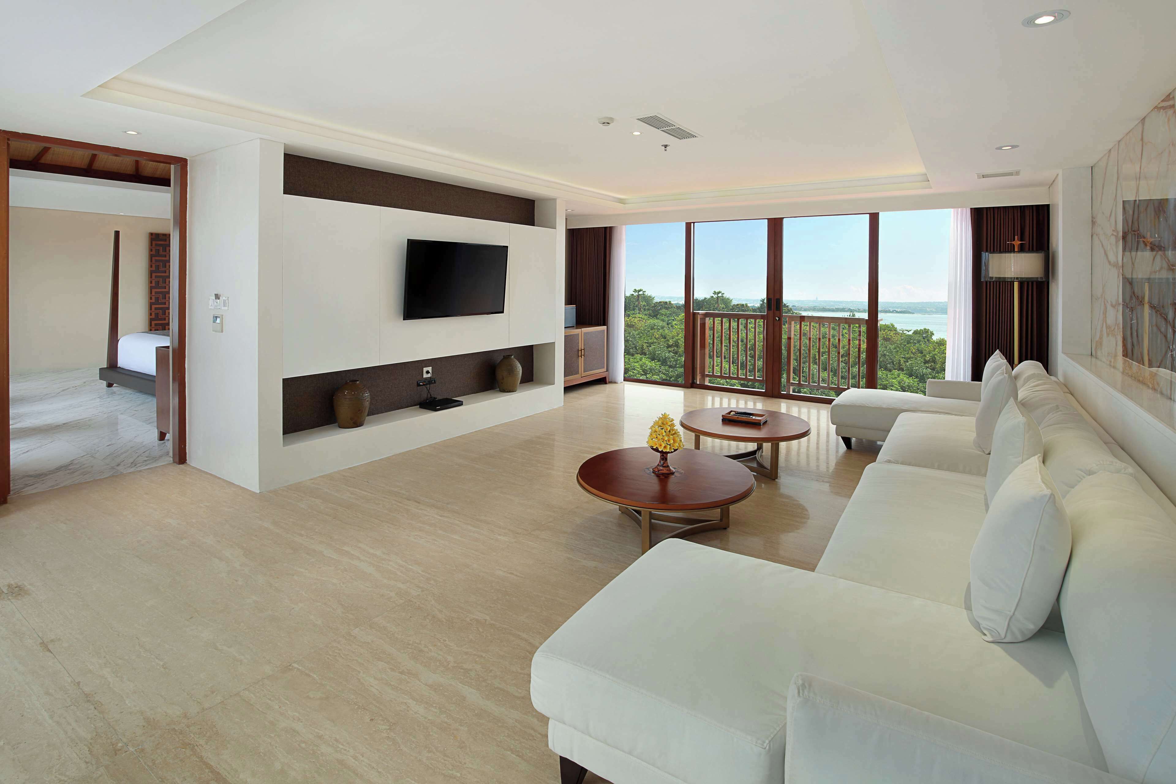 A spacious living room with a large white sofa, a flat-screen TV mounted on the wall, and a balcony with a view of the greenery and water.