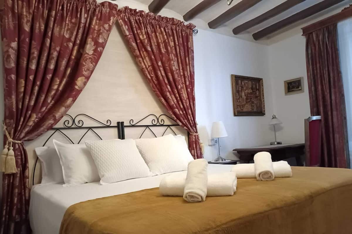 Cama bem arrumada com lençóis brancos e um cobre-leito marrom, cabeceira de madeira e cortinas vermelhas com estampas florais.