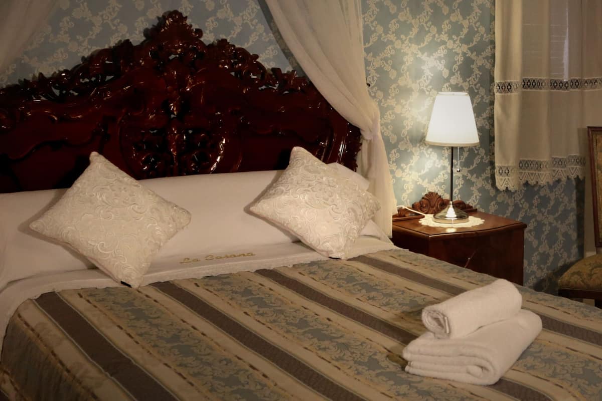 Quarto de hotel com uma cama grande, duas almofadas, uma mesa de cabeceira com abajur, uma cadeira e parede estampada.