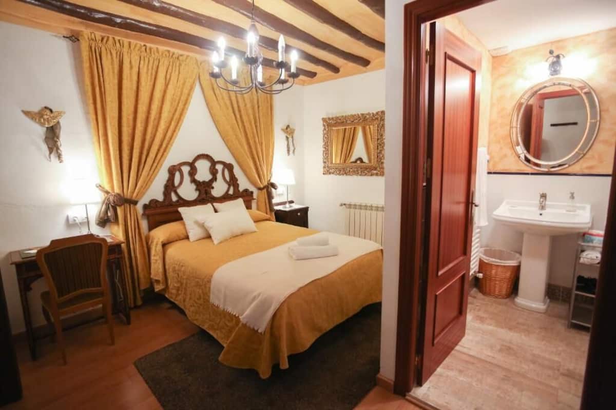 Quarto com cama de madeira, uma escrivaninha, uma cadeira, um candelabro e banheiro com pia e espelho.