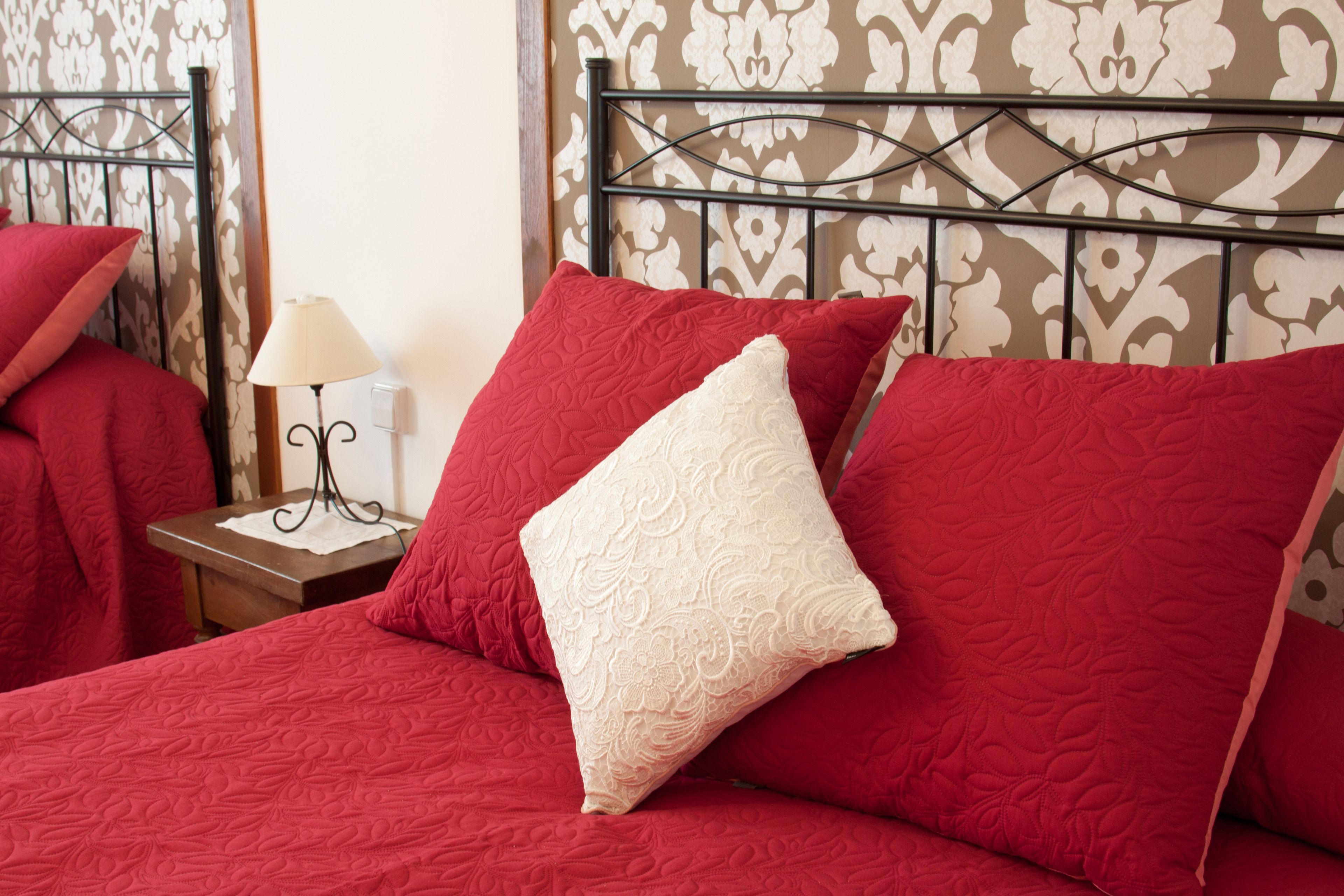 A bed with red bedding and a white pillow, a bedside table with a lamp, and a patterned wallpaper background.