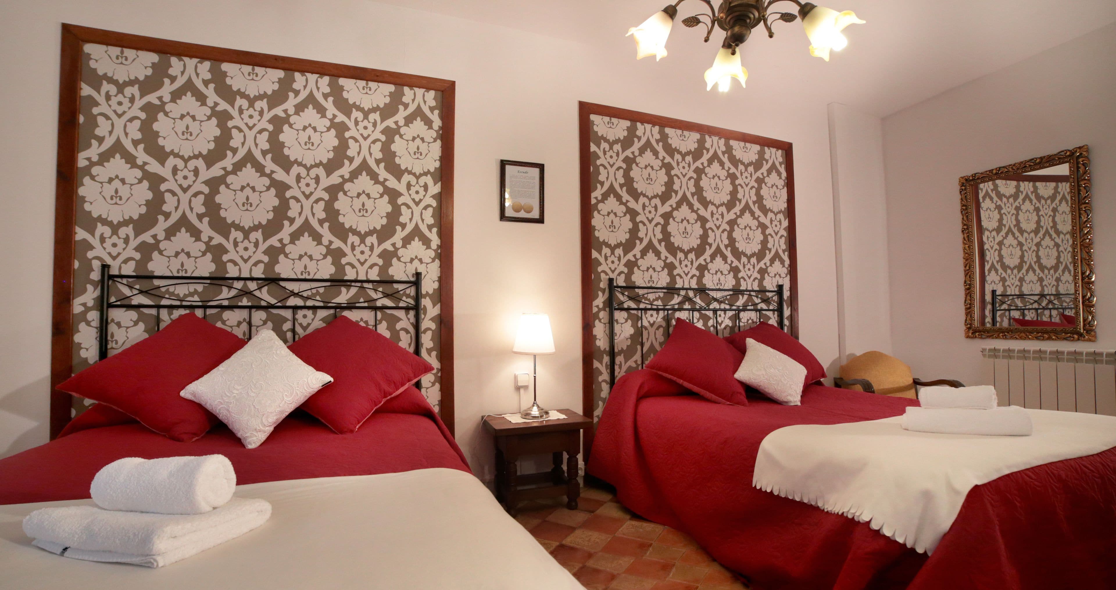 Habitación de hotel con dos camas, cada una con ropa de cama roja y almohadas blancas, una mesita de noche con una lámpara y un espejo en la pared.