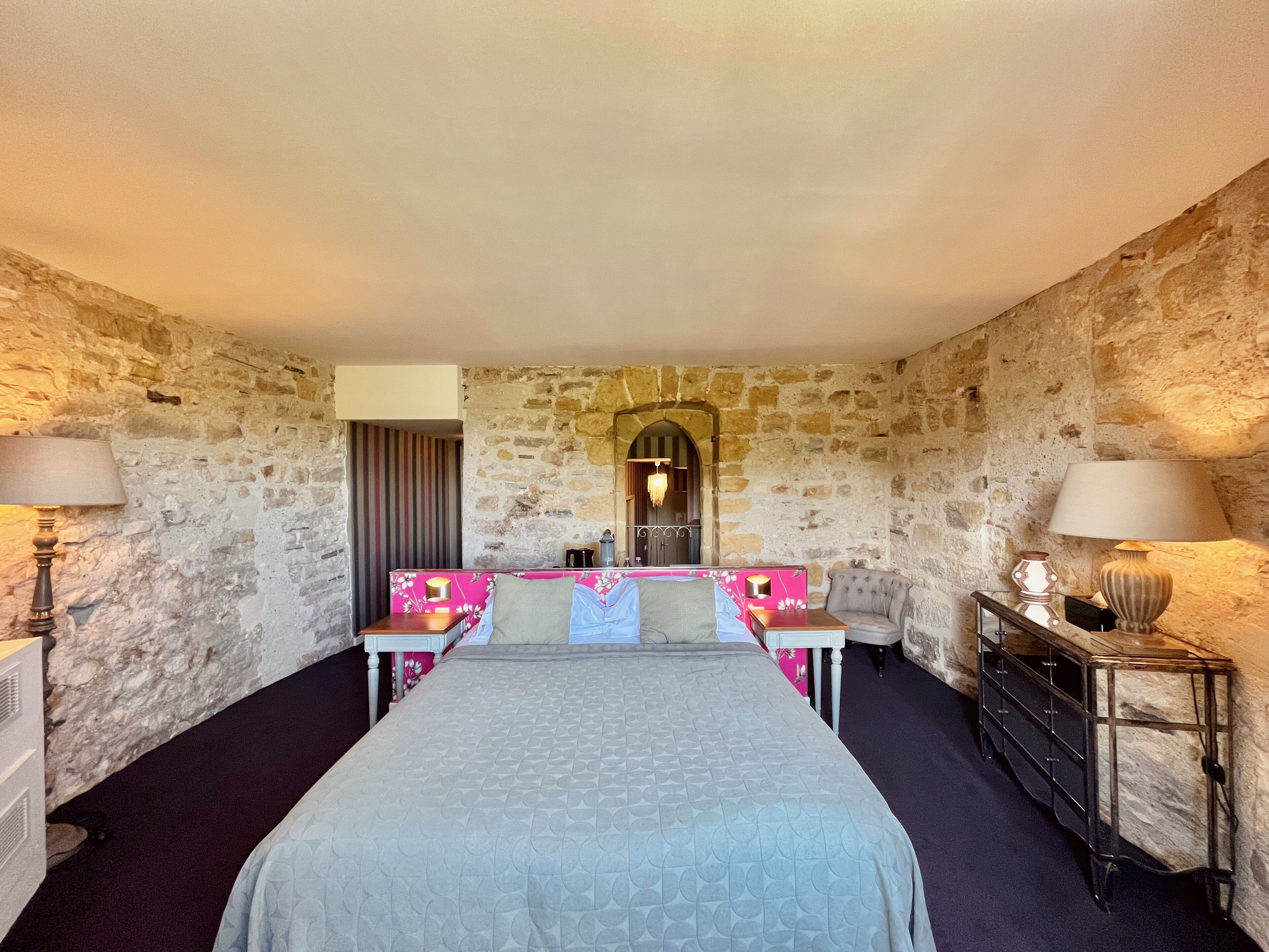 Une chambre d’hôtel avec un mur en pierre, un lit recouvert d’une couverture grise, deux tables de chevet avec des lampes, une commode et un miroir au-dessus du lit.
