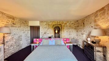 Une chambre d’hôtel avec un mur en pierre, un lit recouvert d’une couverture grise, deux tables de chevet avec des lampes, une commode et un miroir au-dessus du lit.