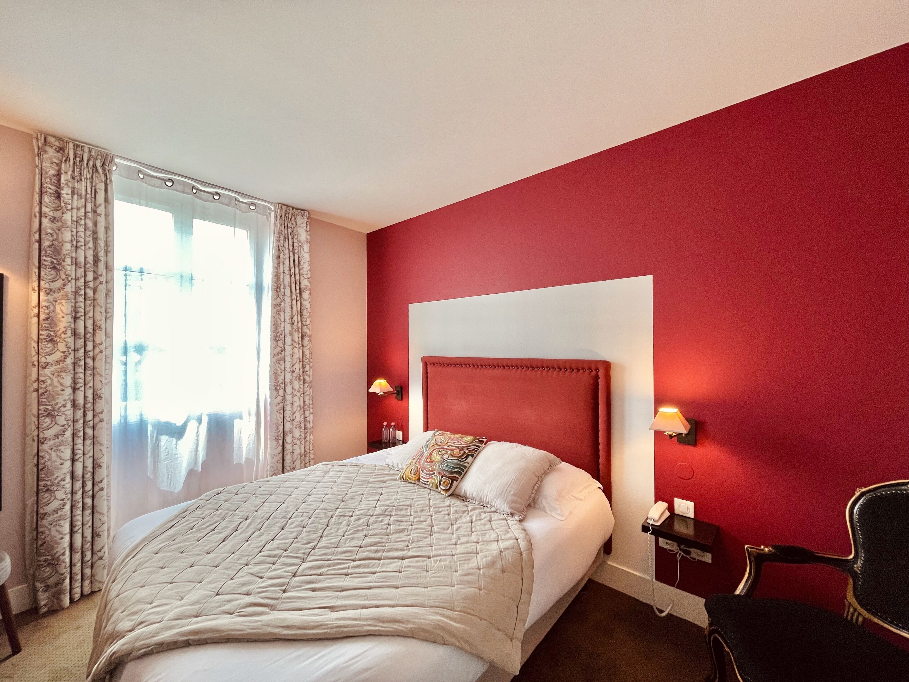A hotel room with a large bed, a red accent wall, and a view of the outdoors through a window with curtains.