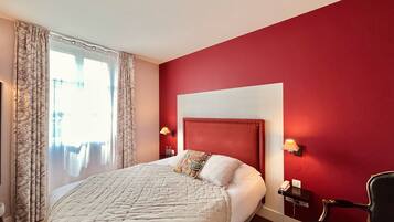 A hotel room with a large bed, a red accent wall, and a view of the outdoors through a window with curtains.