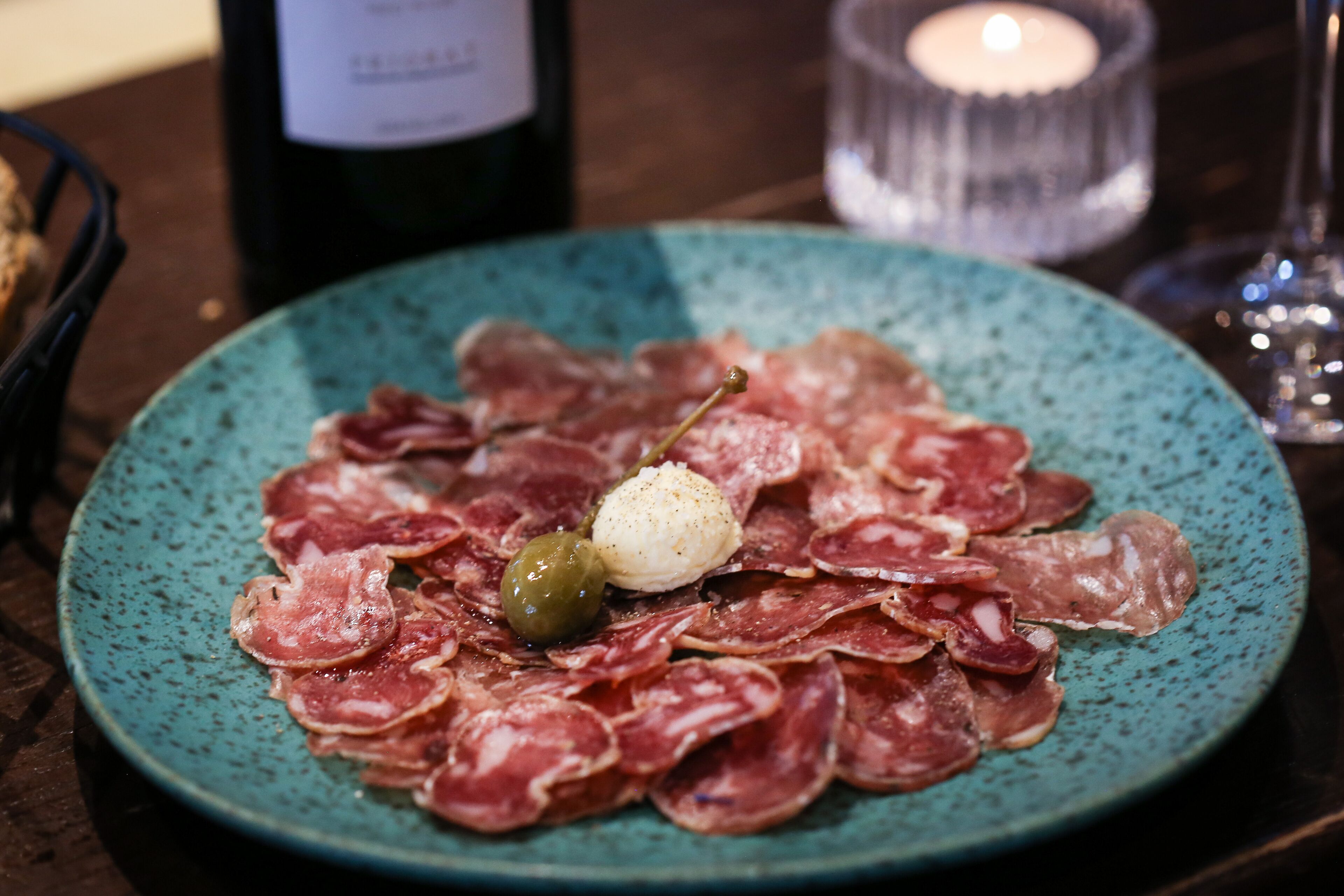Une assiette de charcuteries, accompagnée d’une petite olive et d’une cuillerée de crème, et servie avec une bouteille de vin et une bougie en arrière-plan.