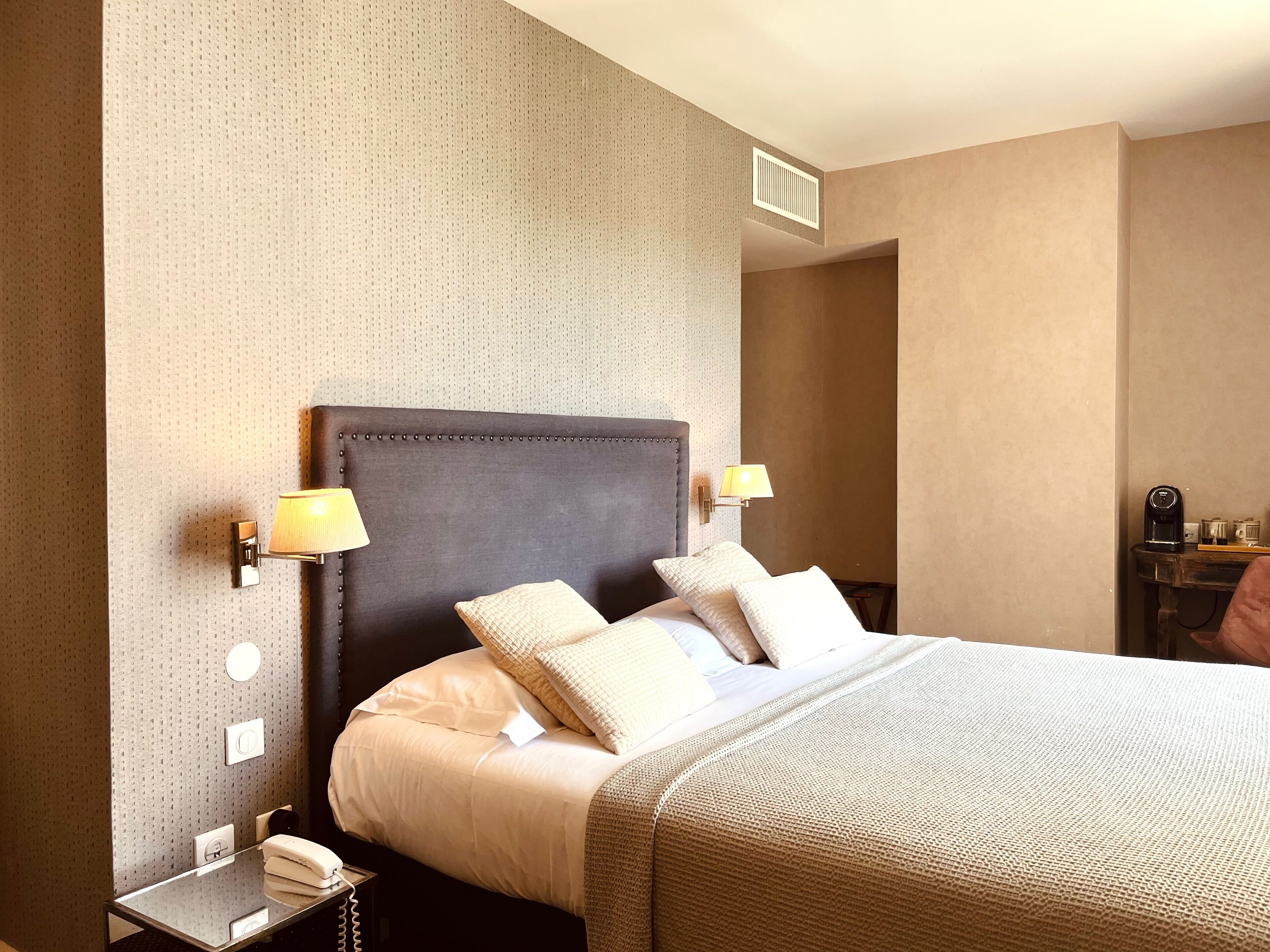 Una habitación de hotel con una cama grande, dos lámparas de noche, una mesita de noche con un teléfono y una pequeña mesa con una cafetera.