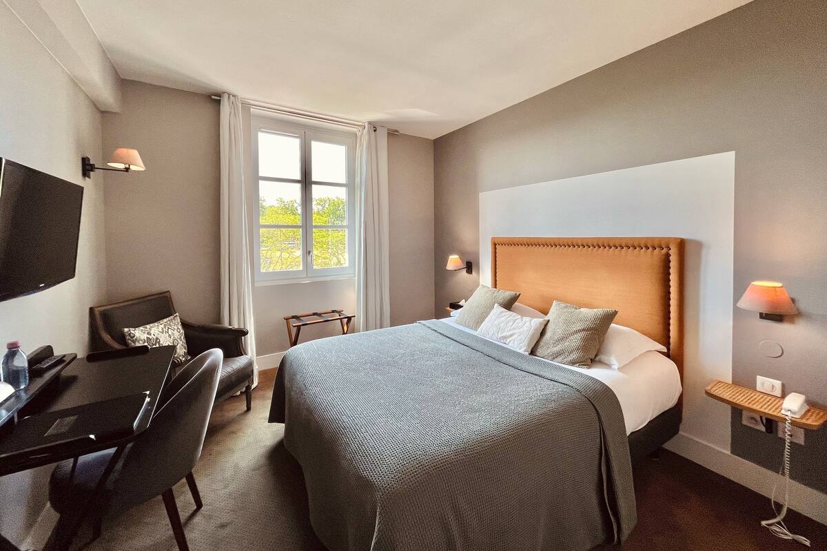 A hotel room with a large bed, a desk with a chair, a TV mounted on the wall, and a window with curtains.