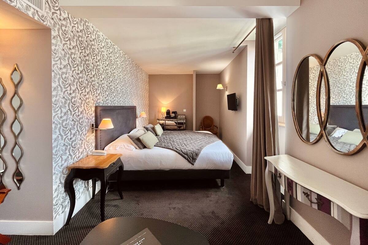 A hotel room with a large bed, bedside tables, a desk, a chair, a TV, and a patterned wall.