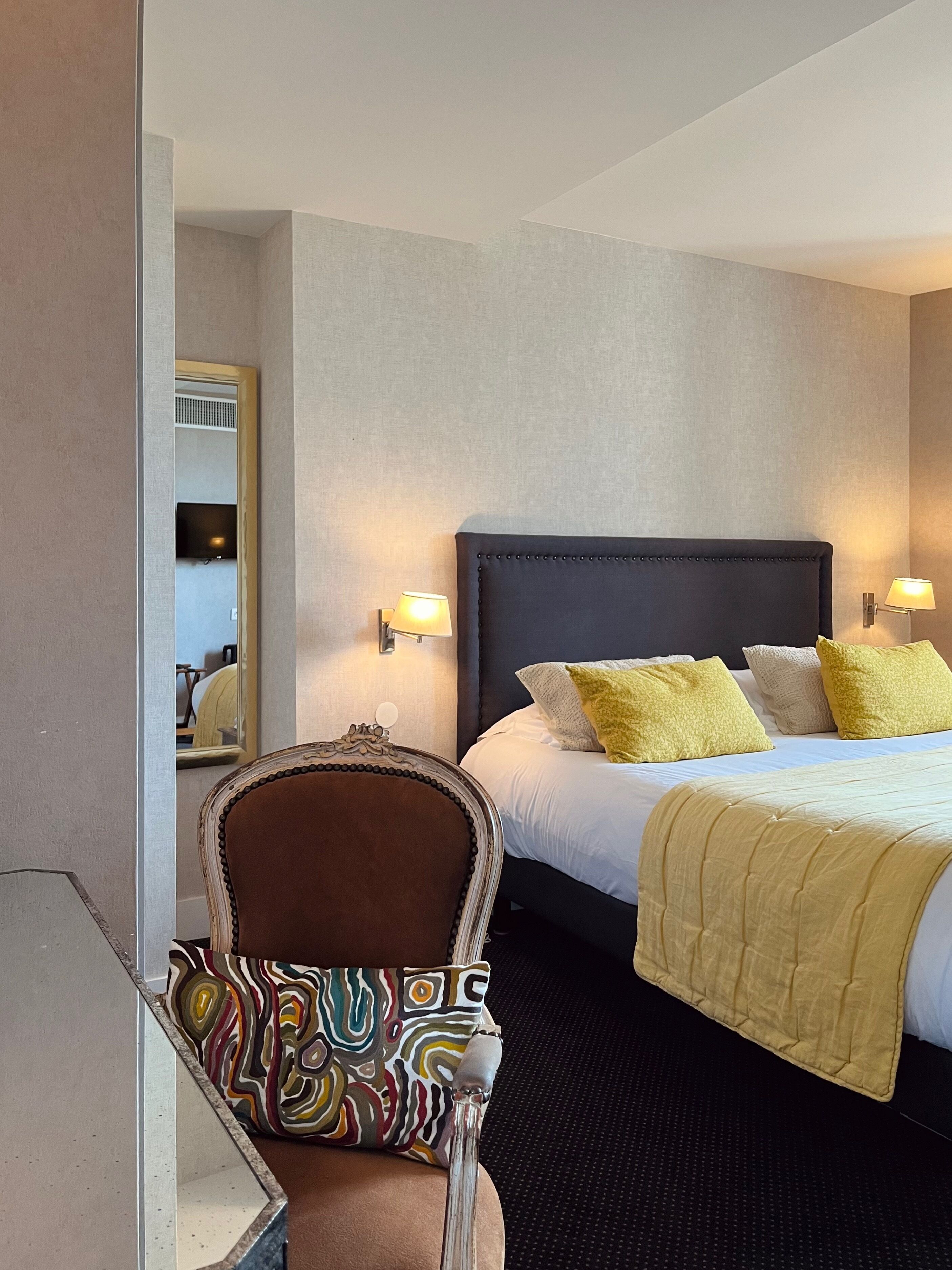 A hotel room with a bed, bedside lamps, a chair with a colorful cushion, and a mirror on the wall.