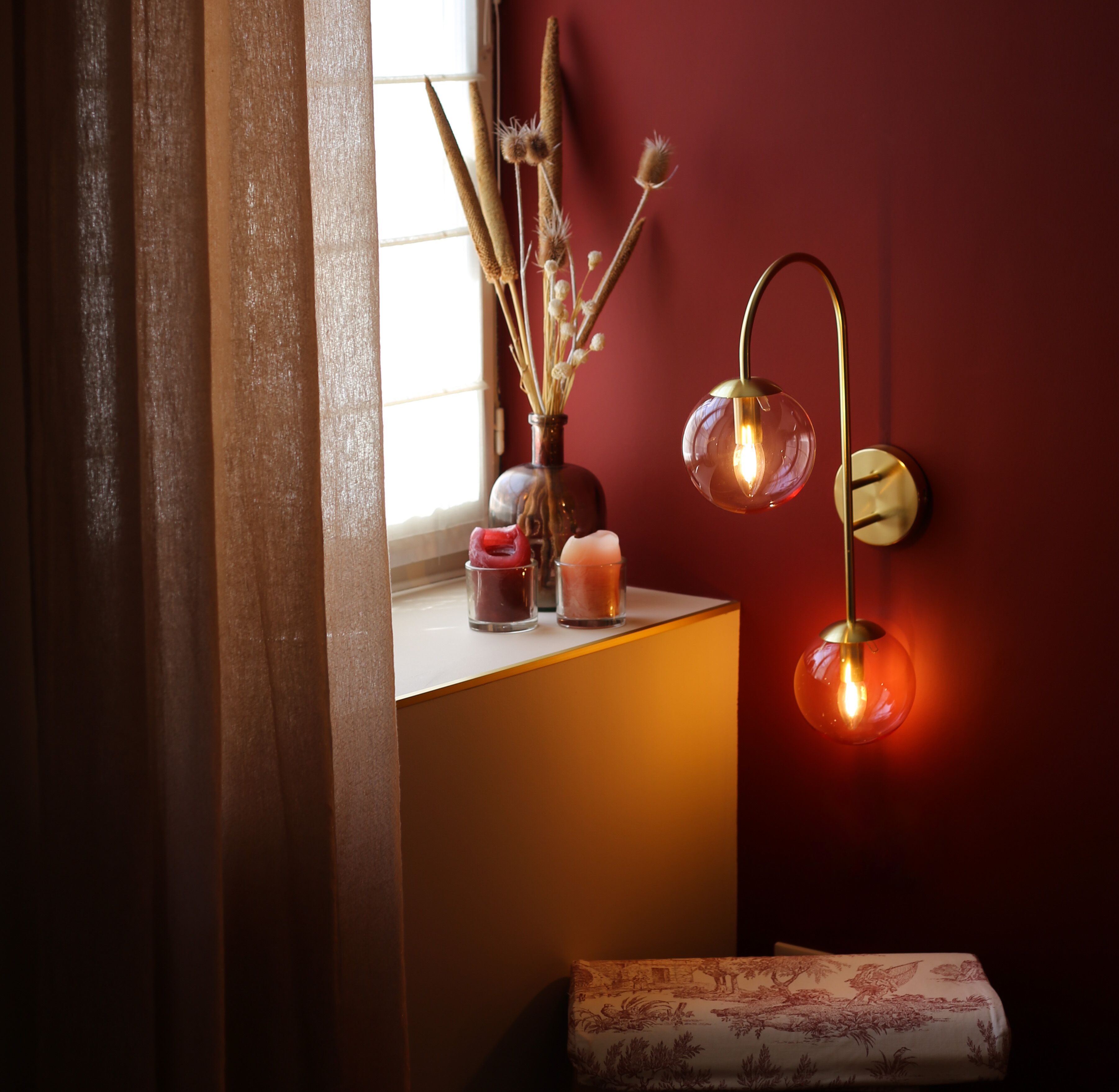 Una habitación con ventana, un florero con plantas secas, dos velas y una lámpara fijada a la pared.