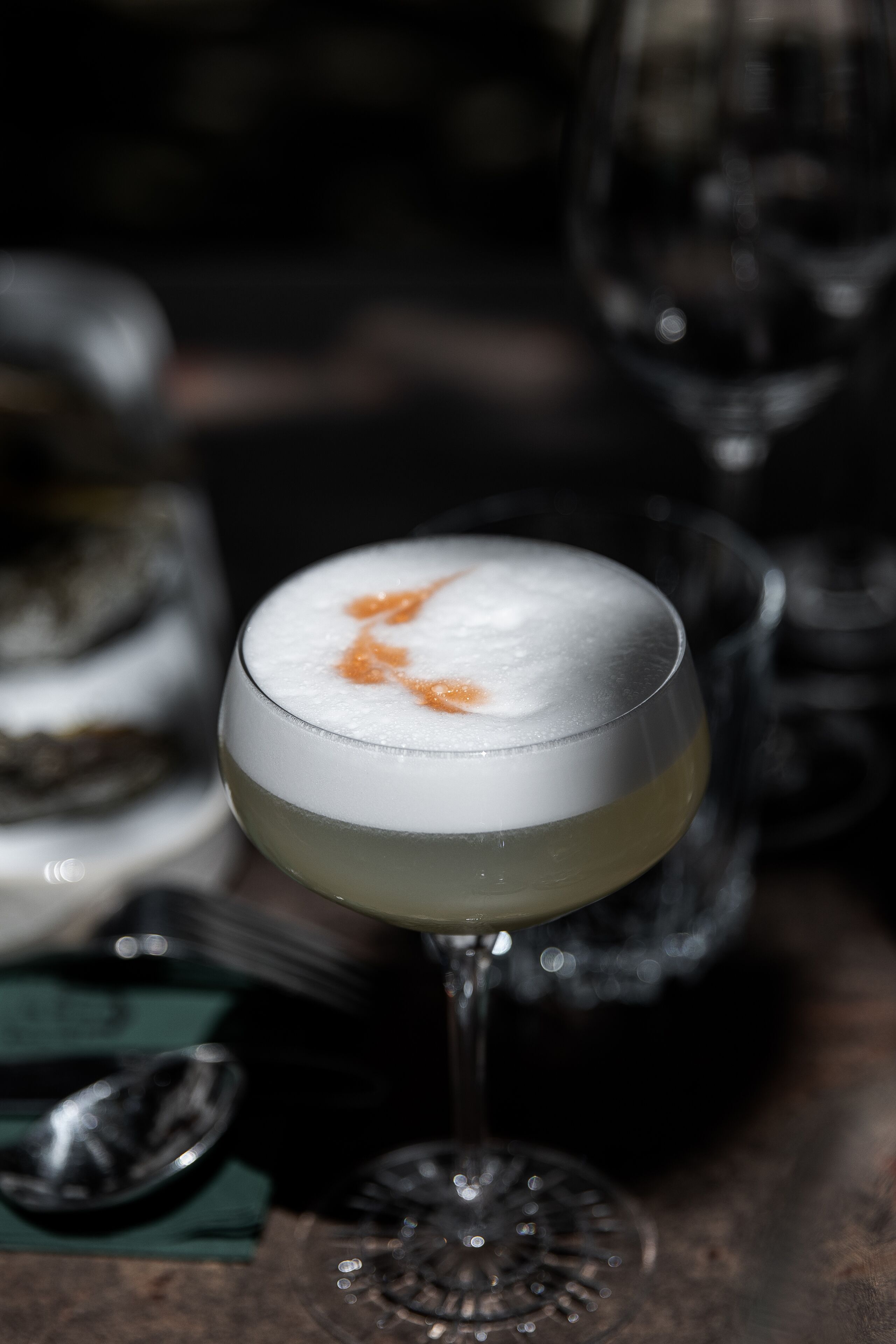A cocktail in a glass with a frothy top, placed on a wooden surface with a spoon and a glass in the background.
