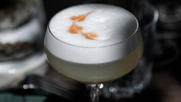 A cocktail in a glass with a frothy top, placed on a wooden surface with a spoon and a glass in the background.