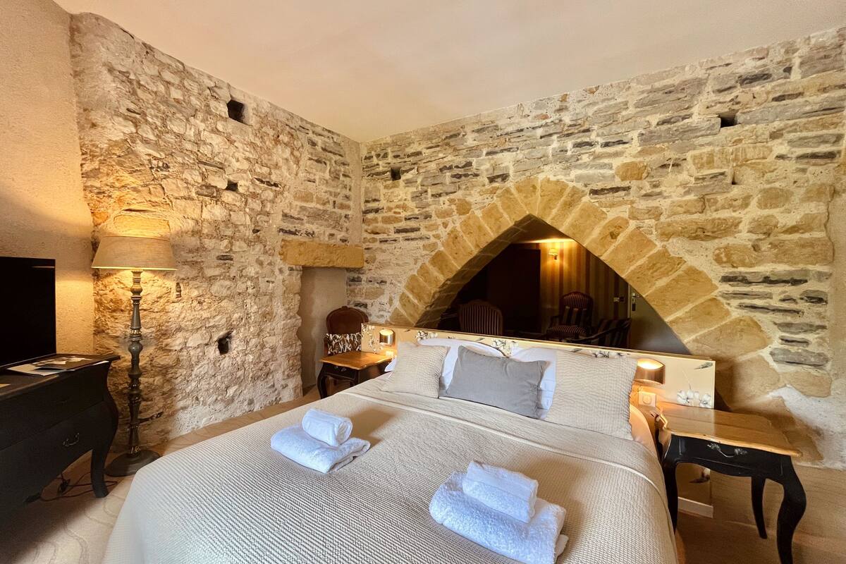 A bedroom with a stone wall, a bed with white linens, a lamp, and a small table.