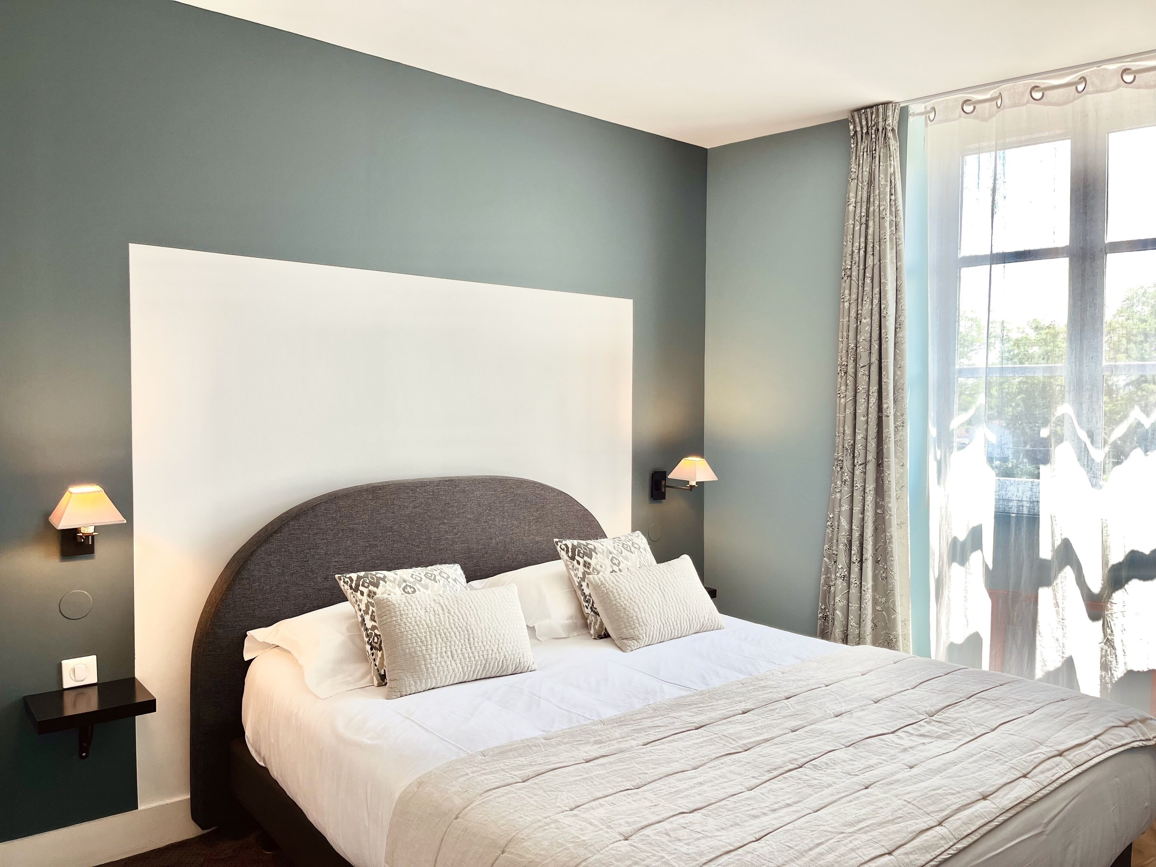Un dormitorio con cama, dos lámparas de noche, una mesita de noche y una ventana con cortinas.