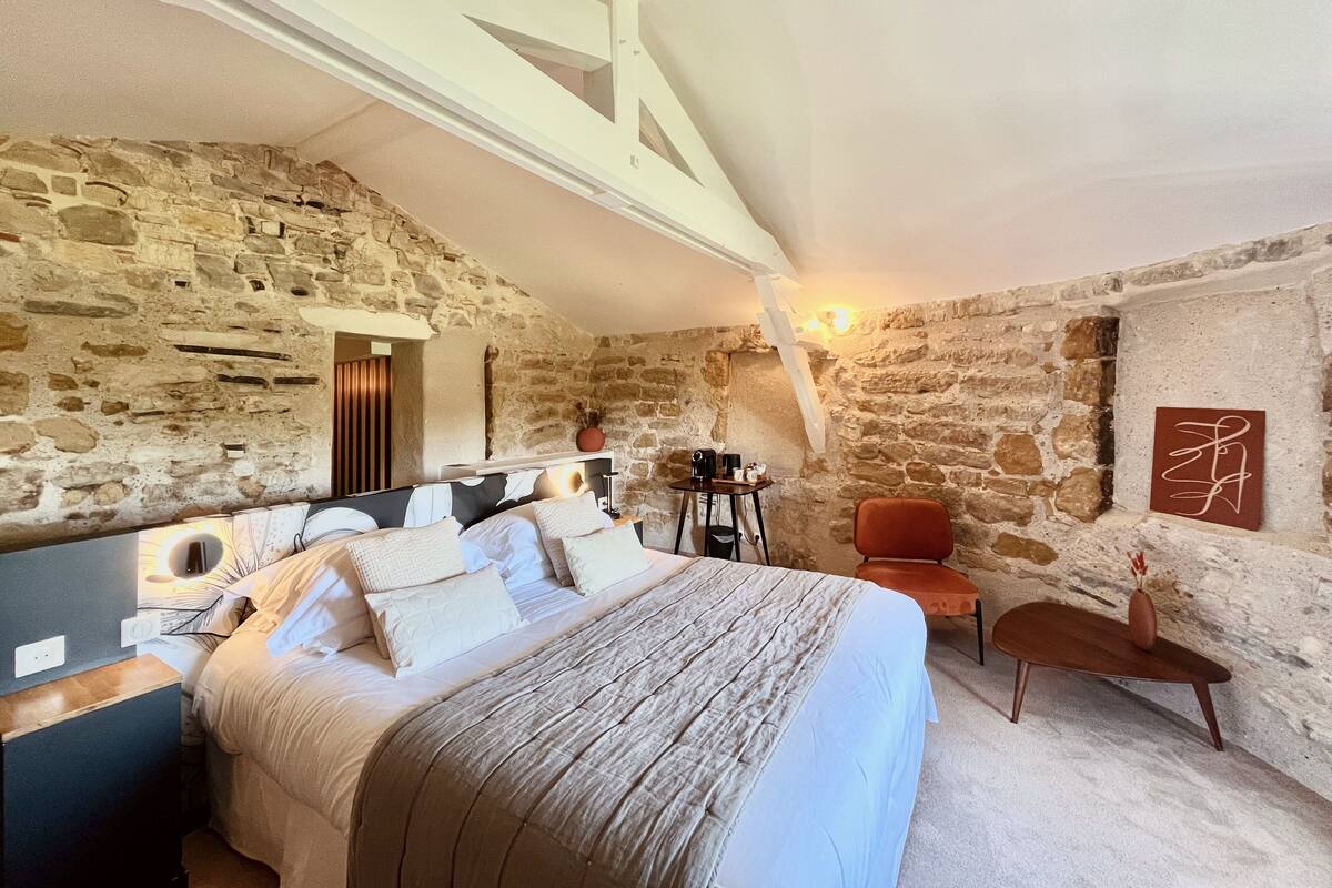 A bedroom with a stone wall, a bed with a headboard, a nightstand, a chair, and a table with a vase.